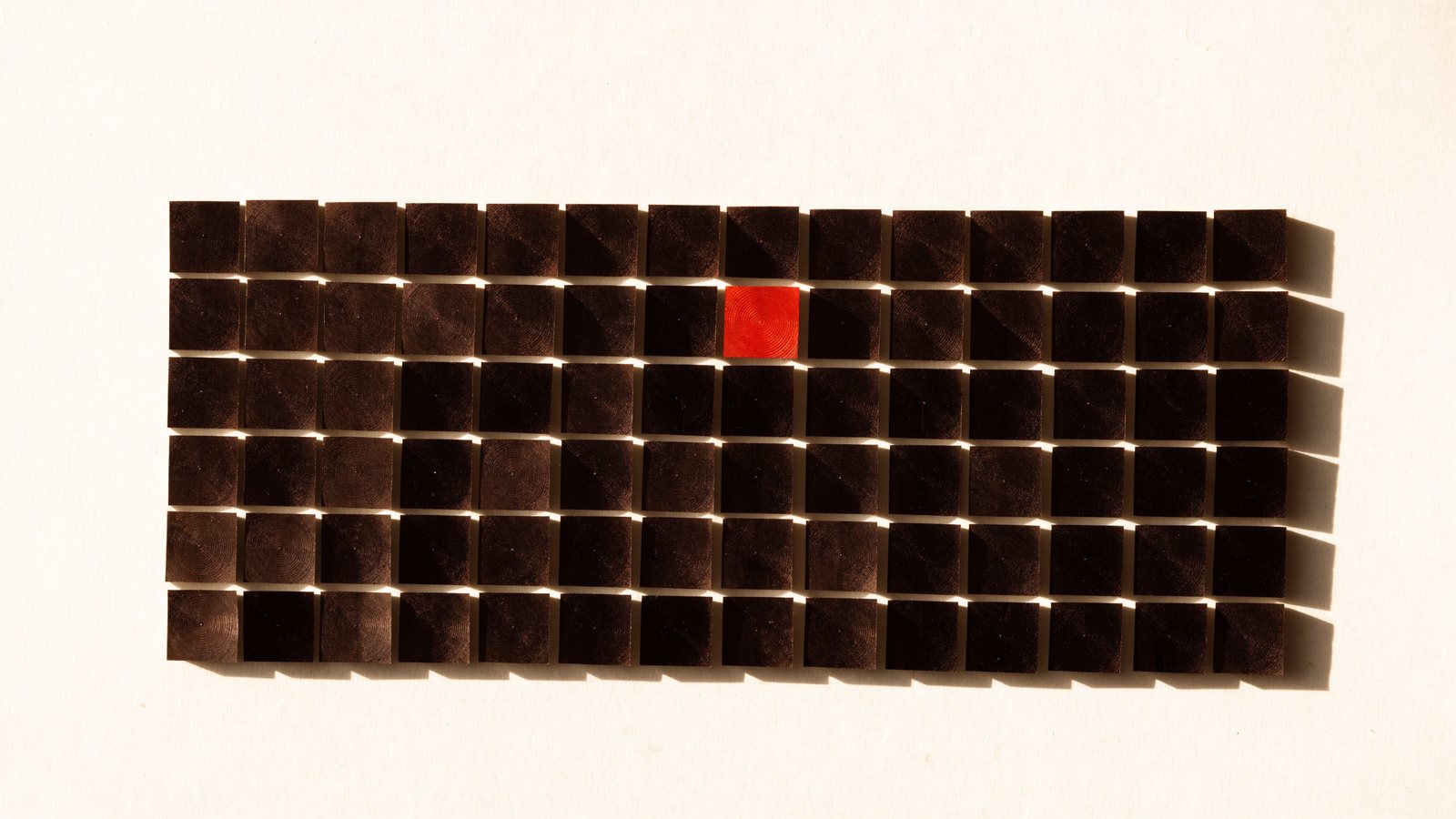 A grid of dark wooden blocks on warm paper, one of them vermilion