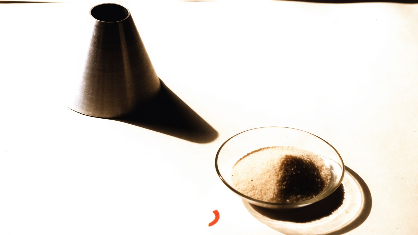 A machined metal cone beside a glass dish of fine sand on warm paper